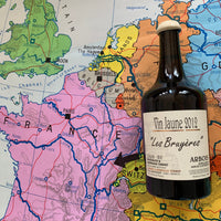 2012 Vin Jaune “Les Bruyéres” fra Bénédicte & Stéphane Tissot i Jura, Frankrig. Lidt tilsat svovl, ved flaskning. Vinflaske med europakort i baggrunden. En pil peger fra flasken, ind på området Jura, i Frankrig.